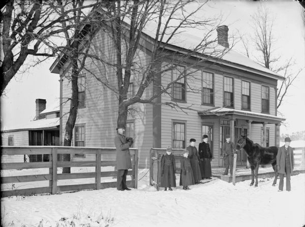 Two men, two women, two girls, and a boy are posing standing in the snow in front of a two-story frame house. The boy is displaying a horse. Possibly the Schuyler Van Gorden home in Hixton, Wisconsin. The man on the left is holding a small animal in his arms.