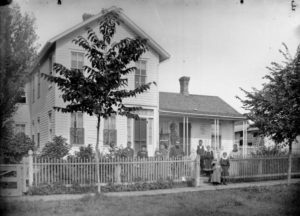 View from road of three men, three women, and three girls posing standing by the gate in a picket fence that is in front of a frame house. One woman is holding an infant. There is a swing on the front porch.