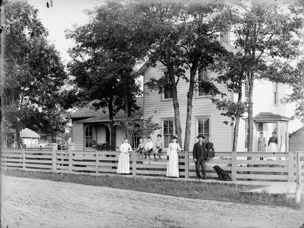 View from road towards two women, three young boys (all barefoot), a young girl and man, and a dog, posing sitting on or standing near a wooden fence along a wooden sidewalk. In the background is a frame house with a young girl and a woman standing on the porch.