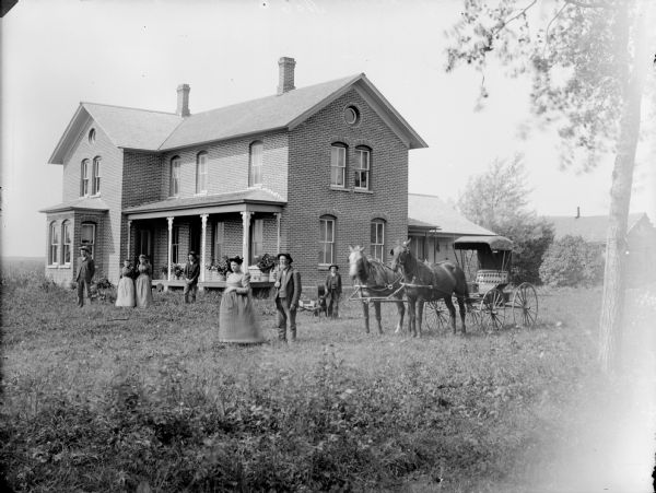 Three men, three women, a boy, and a dog hitched to a small wagon are posed standing in front of a two-story brick house. A team of horses with a buggy is on the right. The woman standing in the center appears to be pregnant.