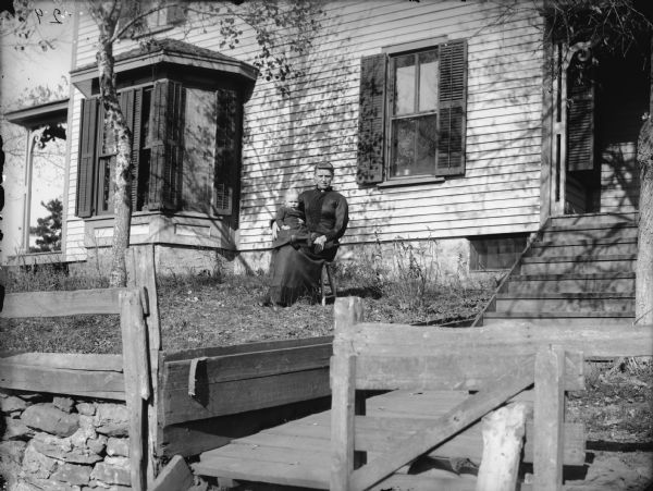 Woman posing sitting on a chair holding a child on her lap in the yard of a frame house.