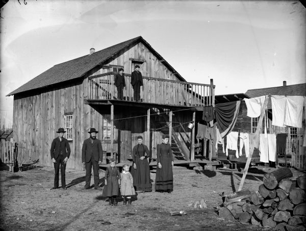 Two women, two men, and two girls standing in front of a wooden house (probably the backyard), near a clothesline with drying wash. Two boys are standing on the edge of second-story porch.
