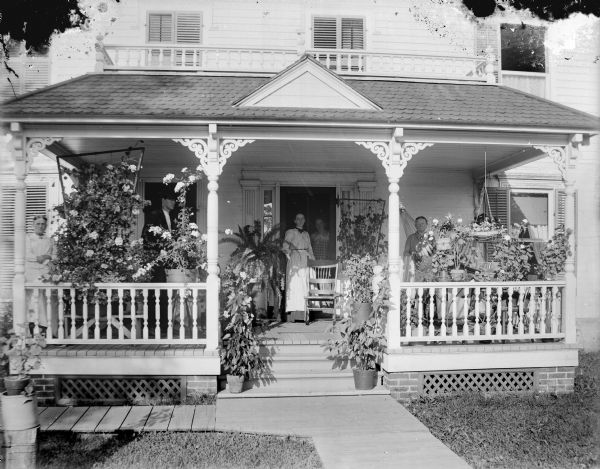 View from sidewalk of a man and three women posed standing on the porch of a frame house among many potted plants. Probably the residence of the Parson family on North Fifth Street. Bill Parson is possibly the man in the dark suit.