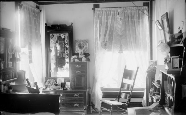Interior of a corner bedroom with numerous keepsakes and photographs, and a dresser, rocking chair, and desk, and a dressing table with mirror. There is a large window on each wall.