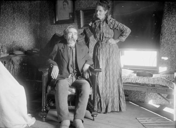 Indoor view of an elderly man posing sitting in a chair, and an elderly woman standing. They are probably in a bedroom. Perhaps the Peterson family.