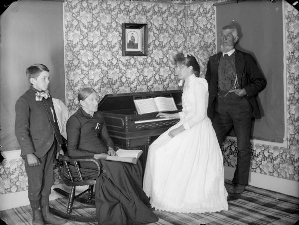 Group of people posing around an organ. A young woman in a white dress is sitting at the organ, facing left, with an older man behind her on the right. On the left, a young boy is standing near an older woman sitting in a rocking chair with a book in her lap.