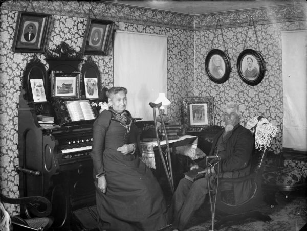 Mr. and Mrs. Simon Overby at home in their parlor with books, family portraits, and an organ with sheet music. Mr. Overby sits in a chair with a cane and a crutch nearby.