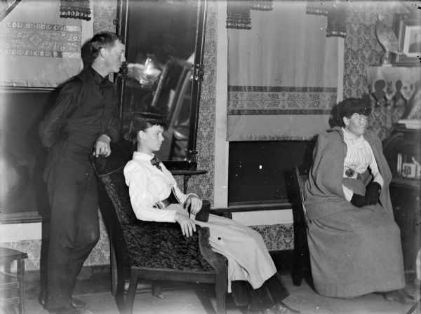 Three people posed in a room with a large mirror between two windows. On the left a man is standing and leaning against the back of an upholstered chair in which a young woman sits. On the right, an older woman wearing a hat a cloak sits in a chair near the window.