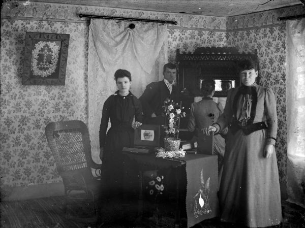 One man and two women are posed standing around a table in a parlor. One of the woman is holding open an album of photographs. Behind them another woman sits at an organ. The reflection of her face can be seen in a mirror above the organ.