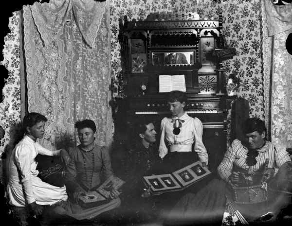 Five women posed sitting on the floor of a parlor in front of an organ holding photograph albums, a man's chest is visible in the organ's mirror, probably Van Schaick himself.