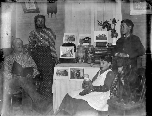 Man and woman pose on left, a girl sitting in a chair is posed holding a doll, with a dog posed sitting in a chair next to her. On the right, a boy is posed standing. Behind the group is a table with a display of photographs, books and objects stored in clear glass jars.