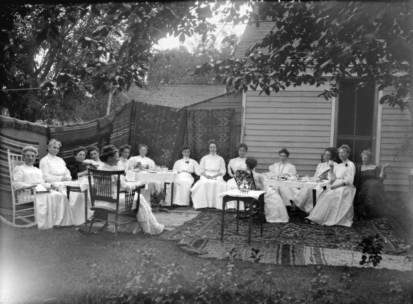 Group of fifteen women gathered around three tables behind home, probably the DMC Club, an exclusive social and sewing society named for an imported brand of French fancywork cotton crocheting, also know locally as the Damn Mean Crowd Club by outsiders. Probably taken at Charles J. Van Schaick's house. Behind them quilts are hanging on a line from the edge of the house all the way across the background, and there are rugs on the ground. Identified by Joanne Dougherty. Mrs. Charles Van Schaick is seated on the far right.
