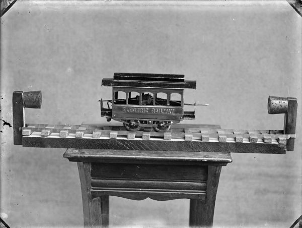 Model train car or street car on a short track. The car is stamped with the words "Electric Railway." The car and track are resting on a small table.