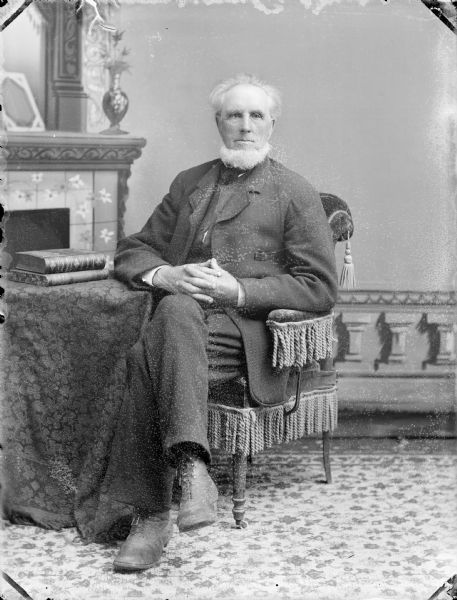 Studio portrait of a man posing sitting in a chair at a table with books on top, possibly Janus (James) Bruce Cartter, 1815-1897. He is in front of a painted backdrop.