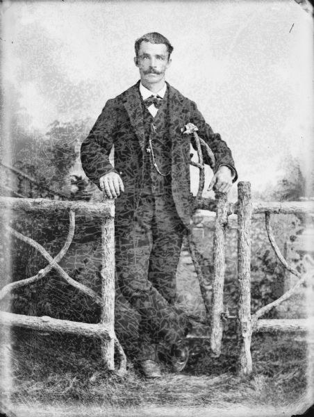 Studio portrait in front of a painted backdrop of a man posed standing in the gate of a wooden fence.	