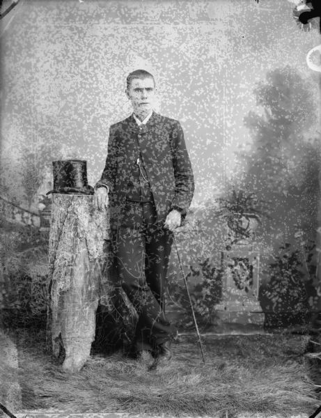 Full-length studio portrait in front of a painted backdrop of a man holding a cane in his left hand, standing beside a tall stump with a hat resting on top of it.