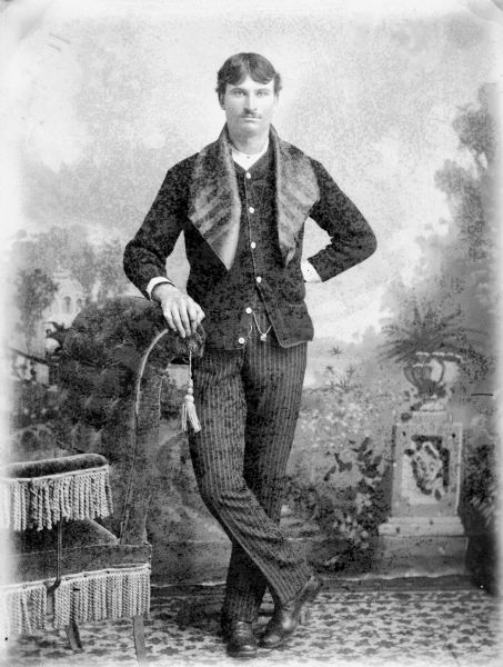 Full-length studio portrait in front of a painted backdrop of a man posed standing and leaning on a stuffed chair.