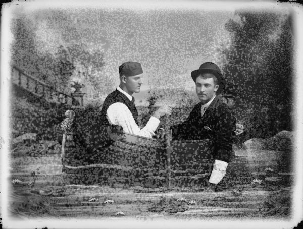 A studio portrait of two men posing sitting in a painted rowboat prop in front of a painted backdrop. The man on the right has his hand in the "water" and is wearing a suit and a hat. The man sitting on the left is wearing a hat and is holding the oars, and has his jacket draped over the edge of the boat.