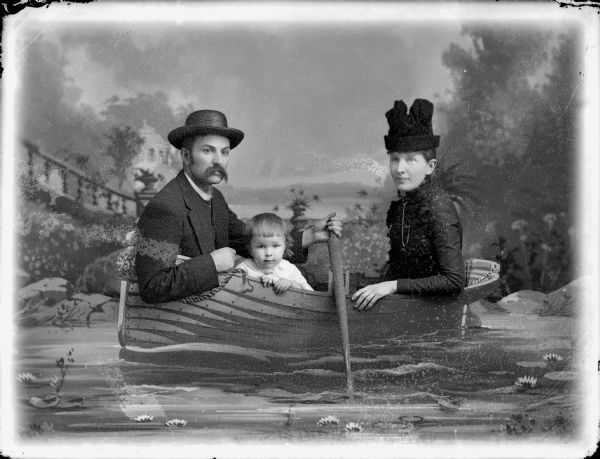 Studio portrait of a man, woman, and child sitting in a rowboat in front of a painted backdrop. Probably Mr. and Mrs. Charles Van Schaick and their son, Shirley.