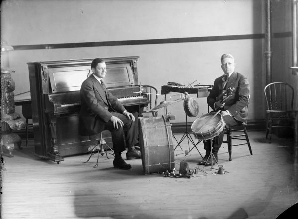 Two men posed sitting next to a piano and drum/percussion set, probably Fortier at the piano and Hoften Hagen sitting at the drums and holding a violin.