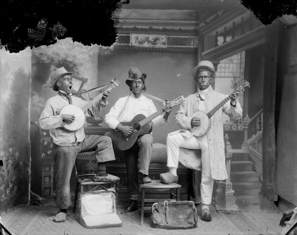Studio portrait in front of a painted backdrop of a woman flanked by two men, all standing, wearing minstrel costumes and blackface makeup.  The men are playing banjos and the woman is playing a guitar. The man on the right is wearing wooden clogs.