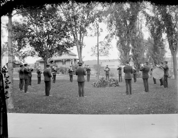 Band posed playing in a circle formation, probably the Alma Center Band, in a concert on the lawn in front of the Jackson County Court House. The house in the background is probably the residence of Jake Spaulding, which later became the place of the Dr. R.C. Gebhardt house.	