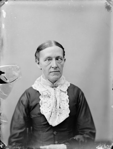 Waist-up studio portrait of an elderly woman and a crossed left eye. She is wearing a lace collar with a pin at the neck.