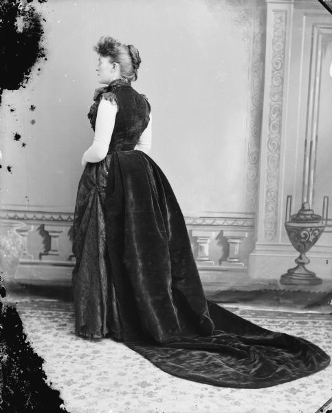 Full-length studio portrait of a woman posed standing sideways with her back to the camera and her head in profile. She is wearing a long black dress. Possibly Mrs. Charles J. Van Schaick.