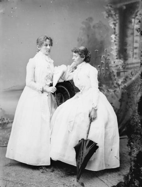 Full-length studio portrait in front of a painted backdrop of a woman posed standing next to a stuffed chair and holding a hand fan, and a woman sitting on the arm of the stuffed chair holding a parasol.