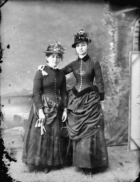 Full-length studio portrait in front of a painted backdrop of two women posing standing. They are wearing dark dresses and hats. One woman is wearing gloves, and the other is holding her gloves.
