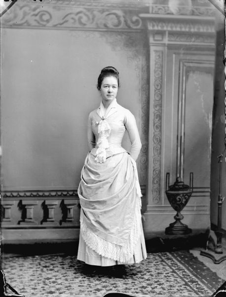 Full-length studio portrait in front of a painted backdrop of a woman posed standing. She is wearing a dress with lace at the collar, and two pearl-like pins with a chain fastened at her bodice.