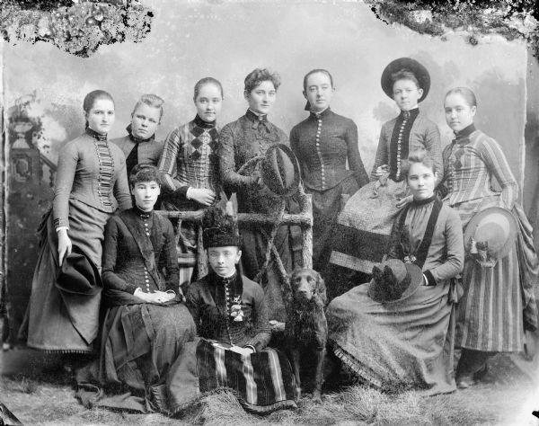 Full-length studio portrait in front of a painted backdrop of nine women, six standing and three sitting, and a dog by a prop wooden fence. The women standing, third from the left, and the last on the right, are the Spaulding twins. Woman on the far left is Alice Mills, and second woman from the right is Julia Ormsby.