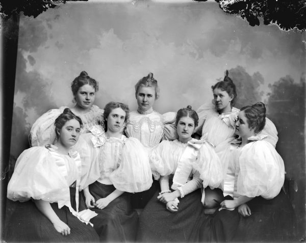 Studio portrait in front of a painted backdrop of seven women, with three standing and four sitting. All the woman have white blouses with puffed sleeves over dark skirts. From left to right, J. Spaulding, unidentified, Alice Mills, and M. Spaulding, and standing from left to right, Julia Ormsby, M.O. Mills, and Sadie Castle.