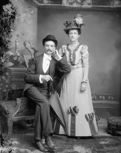 Studio portrait of a man posing sitting on the arm of a chair and holding an umbrella, and a woman posing standing. They are in front of a painted backdrop. Possibly Mr. and Mrs. Olaf Olson. She was the former Mamie Weller.