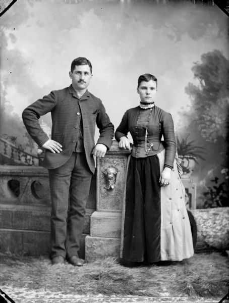 Studio portrait in front of a painted backdrop of a man and woman posed standing next to a prop stone wall.