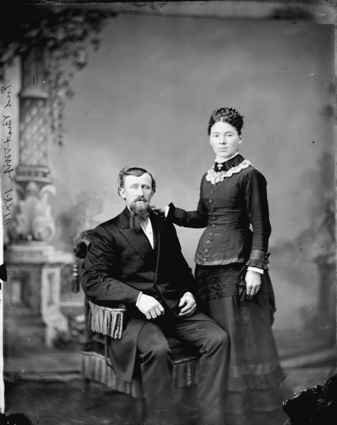 Studio portrait in front of a painted backdrop of a man posing sitting in a stuffed chair and a woman standing, with her arm on his shoulder, facing him with her head turned toward viewer. Probably Mr. & Mrs. Fred Best.