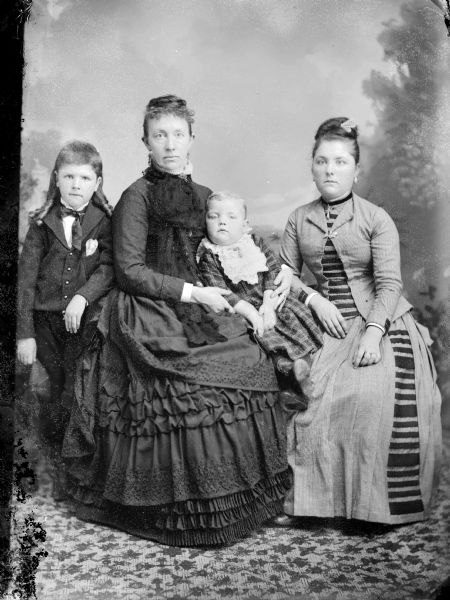 Studio portrait in front of a painted backdrop of two women posed sitting. One woman is holding a child, and another child stands next to her.