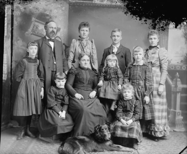 Studio portrait in front of a painted backdrop of three women, five girls, a man, boy and dog.