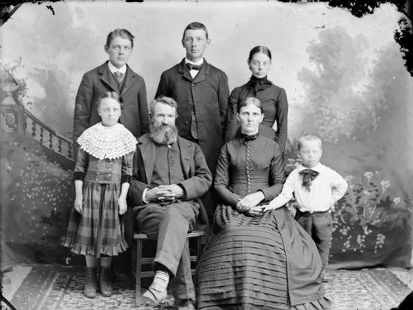 Studio portrait in front of a painted backdrop of a man and woman posed sitting, surrounded by a woman, two young men, a girl and boy.