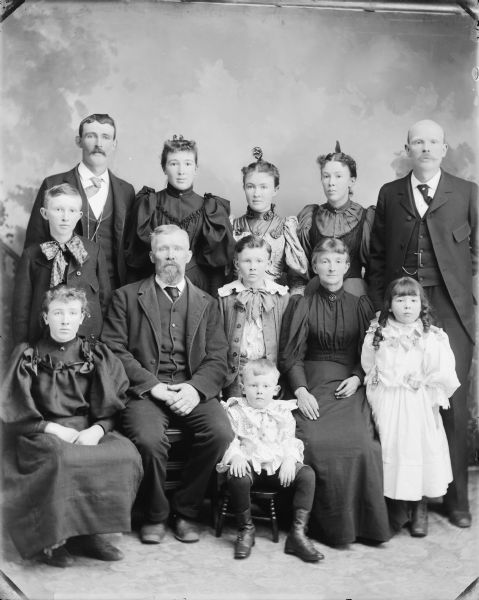 Studio portrait in front of a painted backdrop of a man, woman, boy and girl posed sitting and surrounded by two men, three women, two boys and a girl.