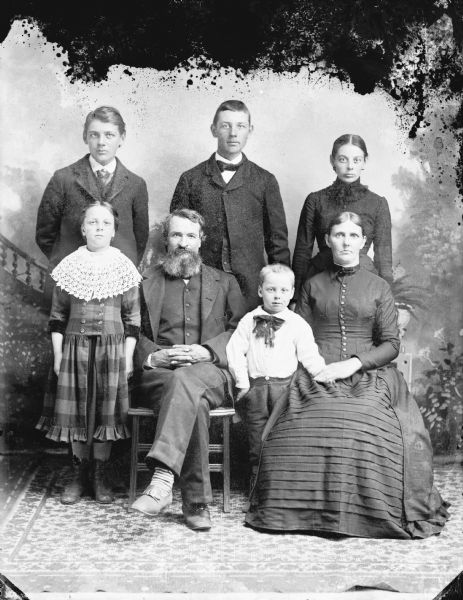 Studio portrait in front of a painted backdrop of a man and woman posing sitting, surrounded by two young men, a young woman, a girl and a boy.