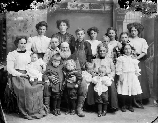 Studio portrait in front of a painted backdrop of a large family, probably the Laib family. Laib was a Civil War veteran and gunsmith and had a shop on Town Creek powered by water. Wilbur "Pat" Laib, wearing the striped shirt at his father's right, fought in World War I. Another son, wearing the overalls, died after he pulled a rifle from a sled. Extreme right is Katherine Waterman, later Mrs. Lud Johnson. Second standing woman from the left is probably Mrs. S.E. Voss.