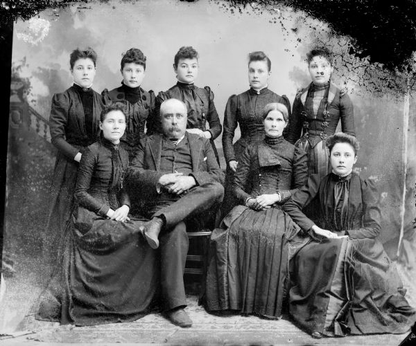 Studio portrait in front of a painted backdrop of eight women, three of whom are sitting, and one seated man.