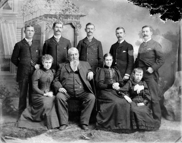Studio portrait in front of a painted backdrop of an elderly man and woman posing sitting surrounded by two seated young girls and five men, probably the H.B. Mills family. Standing, from left to right, Hugh, Edward, Alex, John, and Thomas. Sitting, from left to right, Mary, H.B., wife/mother, and Margie.