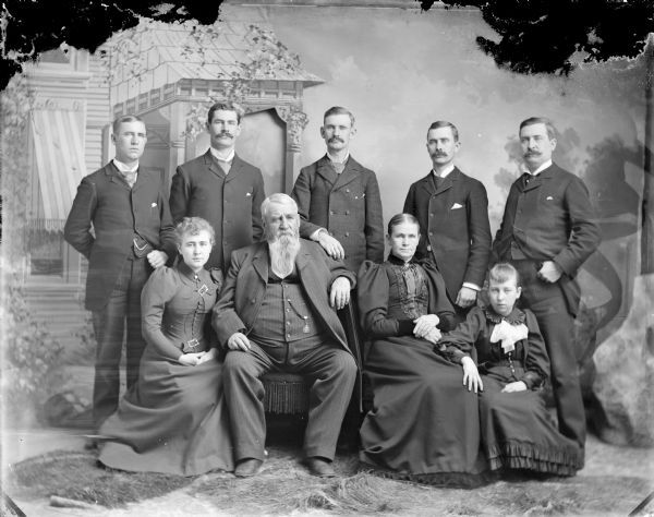 Studio portrait in front of a painted backdrop of an elderly man and woman posing sitting surrounded two seated young girls and five men, probably the H.B. Mills family. Standing, from left to right, Hugh, Edward, Alex, John, and Thomas. Sitting, from left to right, Mary, H.B., wife/mother, and Margie.