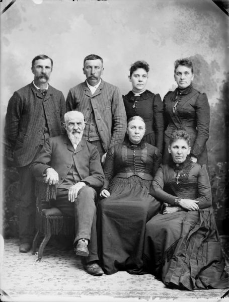 Studio portrait in front of a painted backdrop of an elderly man and two women posed sitting in front of two standing men and two standing women.	