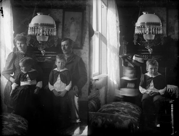 Two exposures of interior portraits. Left is a seated woman holding on to smaller boy both looking left, with seated man and standing older boy facing forward. On right, from almost the same position, are the two boys, with the older boy in the same position, the younger still seated but in motion and barely visible.
