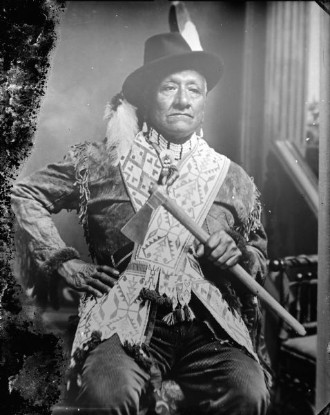 Studio portrait in front of a painted backdrop of a Native American man posed sitting. Henry Rice Hill, wearing native dress, and holding a steel head tomahawk.	