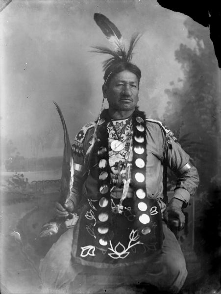 Studio portrait in front of a painted backdrop of a Native American man posing sitting and wearing native dress, probably Thomas Thunder.