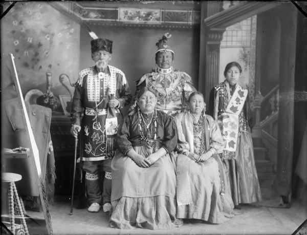 A studio portrait in front of a painted backdrop of Winnebago Indians: John Hazen Hill, Alex Lonetree, daughter of Alex Lonetree, Mrs. Alex Lonetree, and Wolf Woman.